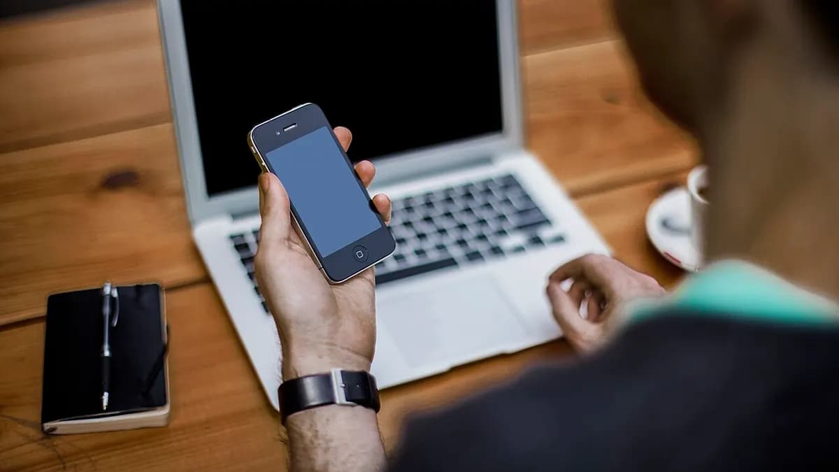 A person holding a smartphone above an open laptop on a wooden table